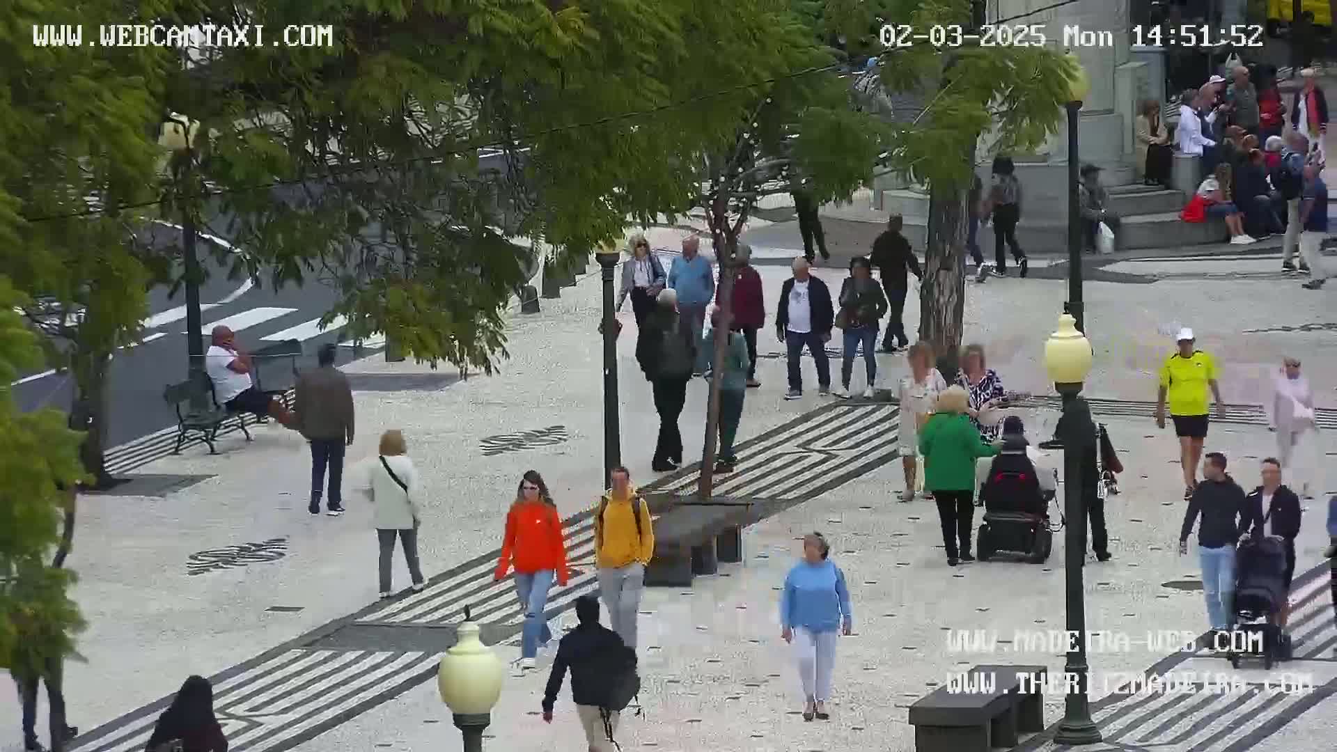 A mostly sunny outdoor plaza with many pedestrians walking on a patterned stone walkway, some sitting on benches, and a person in a wheelchair.