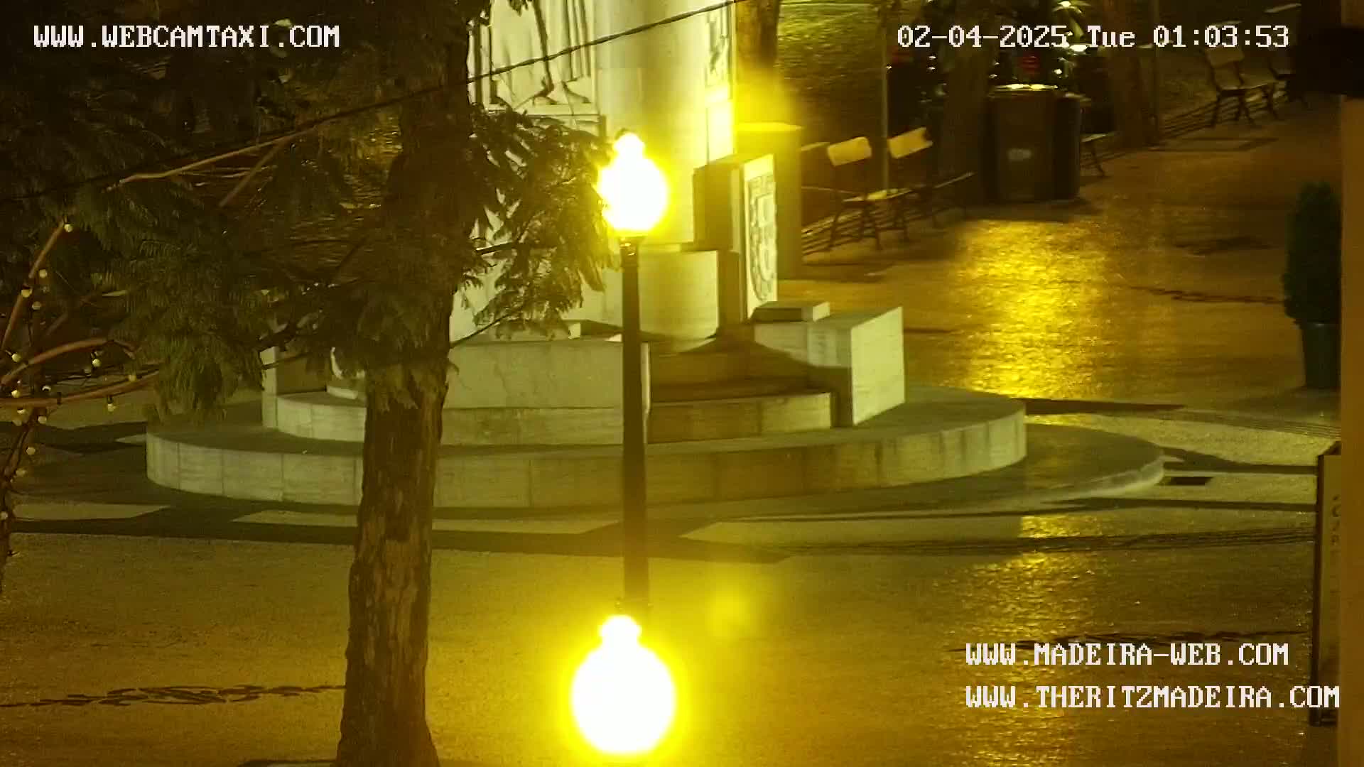 A nighttime, wet scene shows a stone monument with a lamppost in front, partially obscured by a tree.