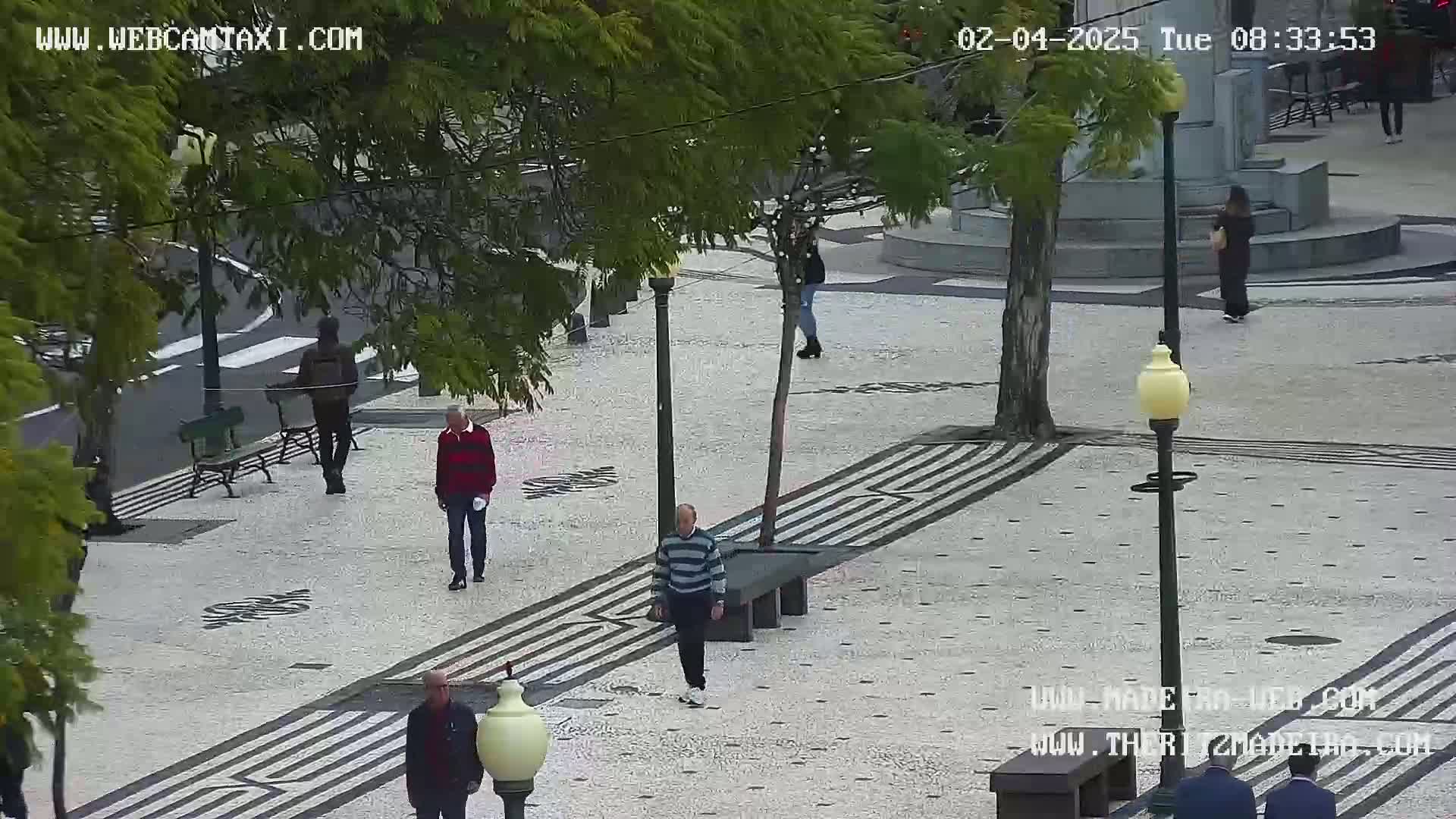 A paved plaza with several pedestrians walking past benches and lampposts on an overcast day.