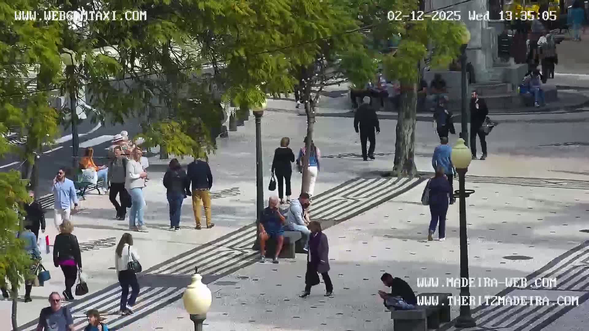 A paved plaza with many pedestrians, some sitting on benches, under a sunny sky, surrounded by leafy trees.