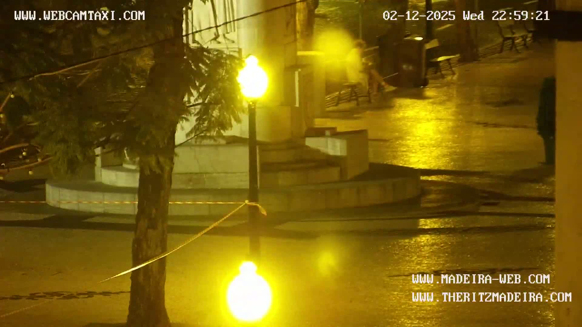 A wet, nighttime outdoor scene shows a person walking near a lamppost and a tree in a plaza-like area.