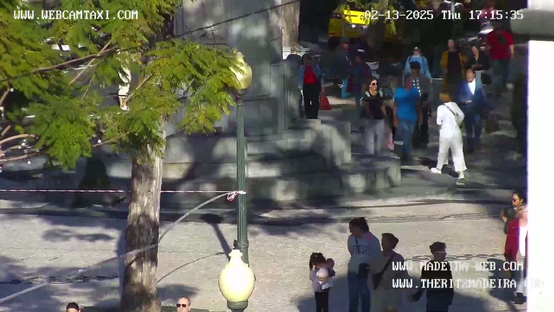 A sunny outdoor scene shows a paved plaza with several people walking around a monument, some standing in small groups, near a lamppost and tree.