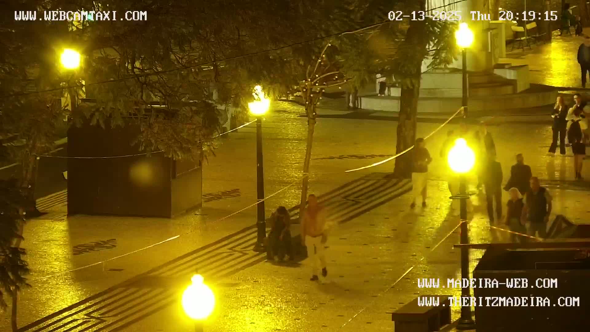 A nighttime, rain-slicked plaza is illuminated by numerous streetlights, and several people are walking through the area.