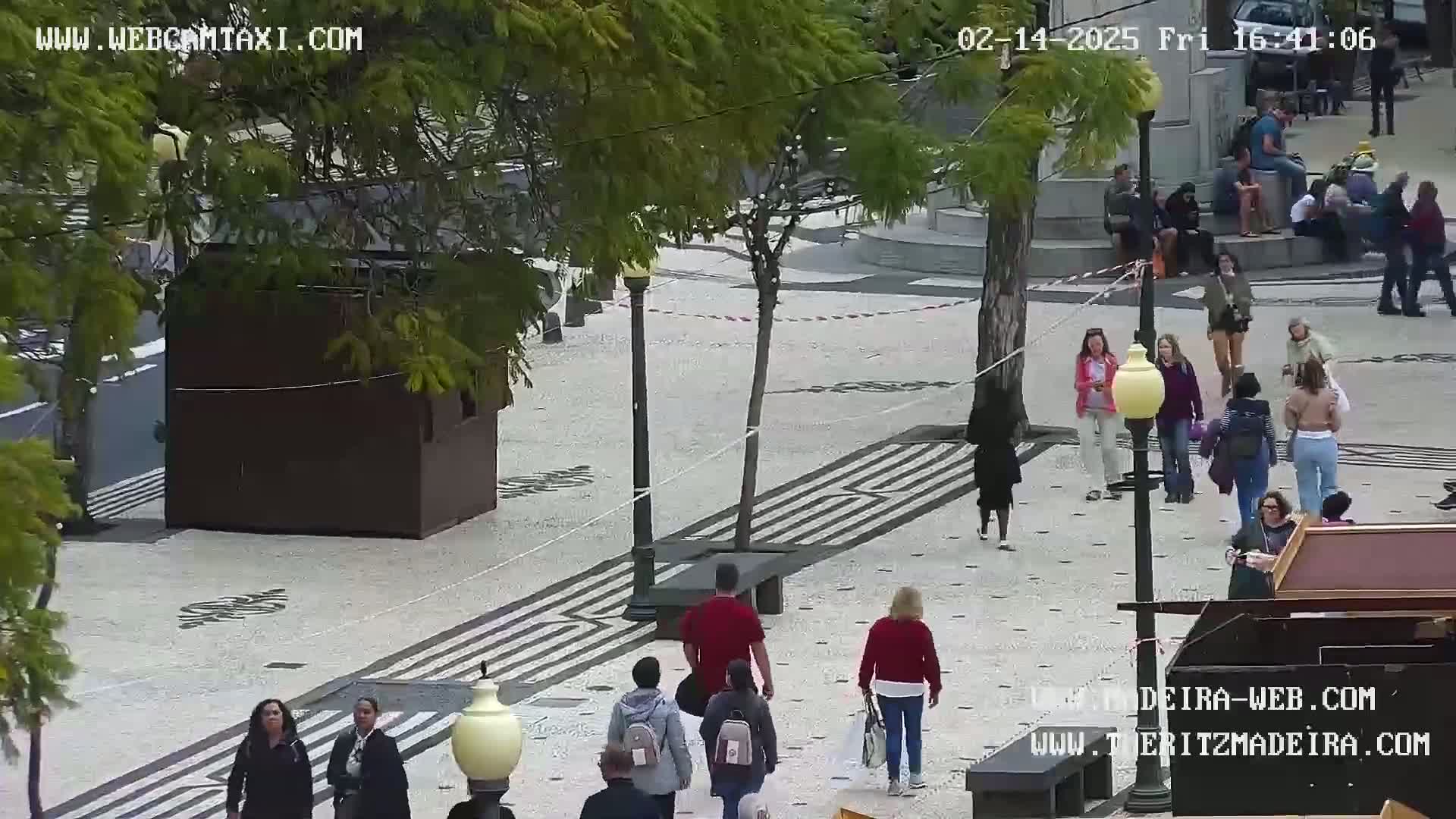 A mostly sunny outdoor plaza, paved with patterned stone, features several people walking among trees, benches, and small kiosks.