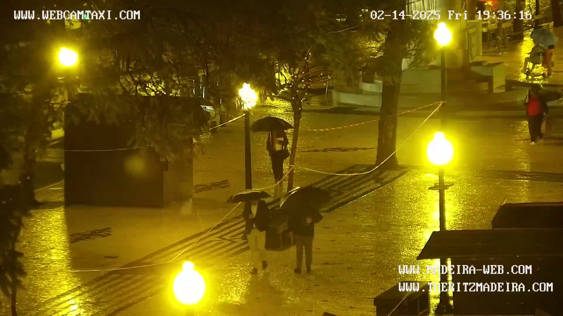 A rain-slicked nighttime square is illuminated by bright yellow streetlights, with several people carrying umbrellas walking through the wet pavement.