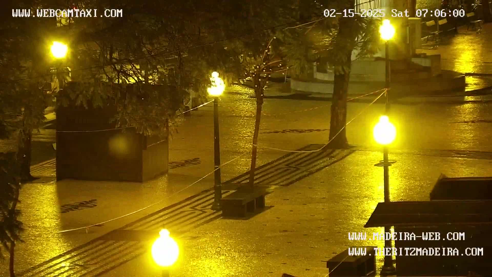 A rain-soaked plaza at night is illuminated by numerous streetlights, reflecting the yellow glow onto the wet pavement.