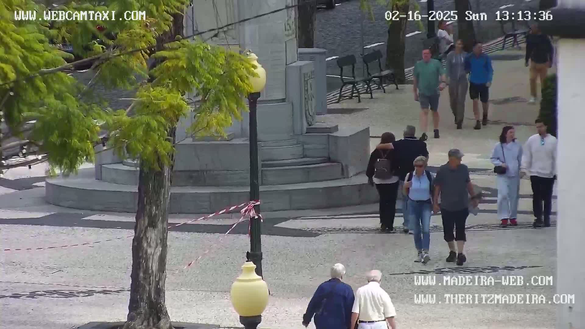 A sunny outdoor plaza scene shows several groups of pedestrians walking on a stone pavement around a monument, with a tree to the left.