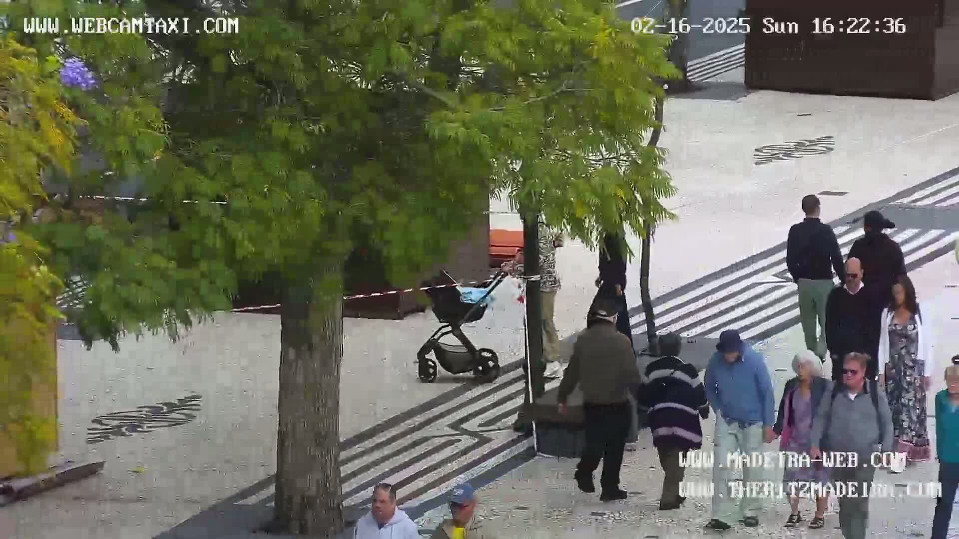 A paved outdoor plaza with several people walking around, including a person pushing a baby stroller, under a partly sunny sky.