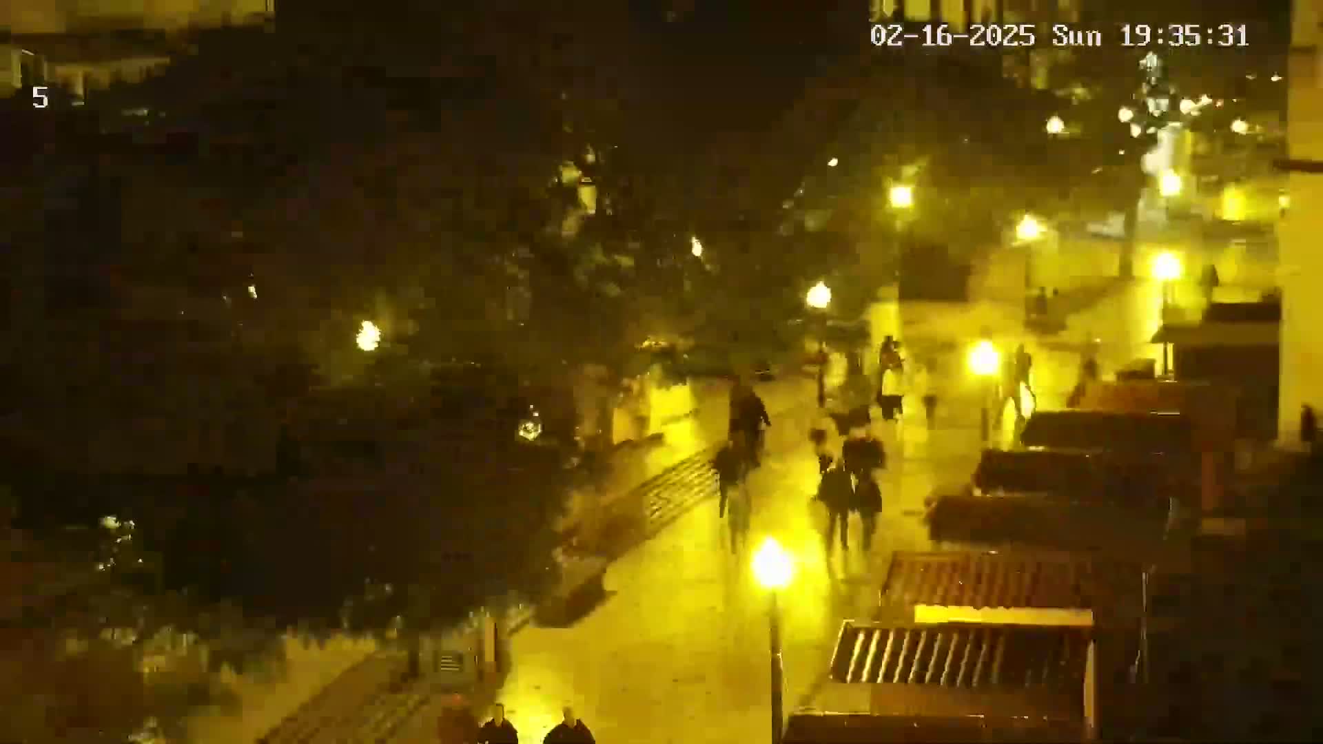 A nighttime, rain-slicked street scene shows several people walking past buildings and under streetlights.