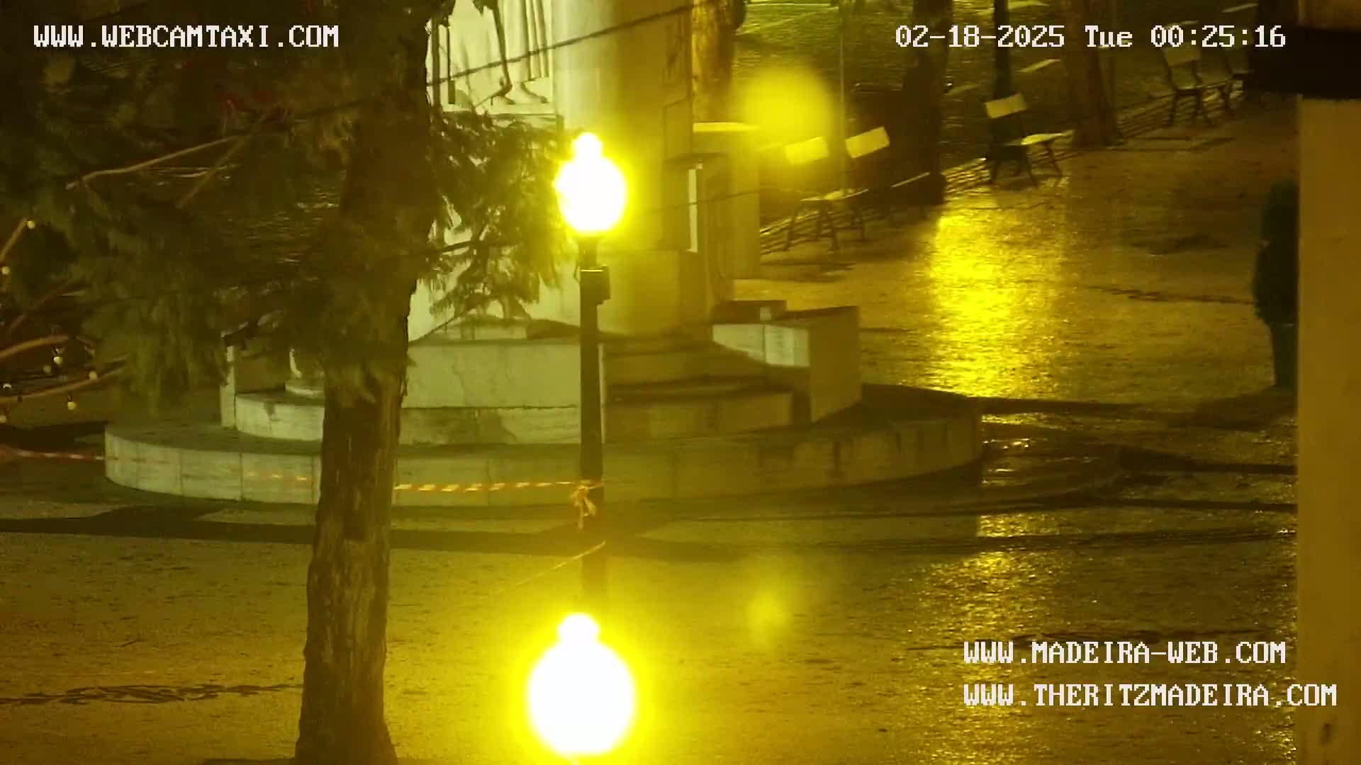 A wet, nighttime scene shows a plaza with benches and a lamppost, illuminated by yellow streetlights.