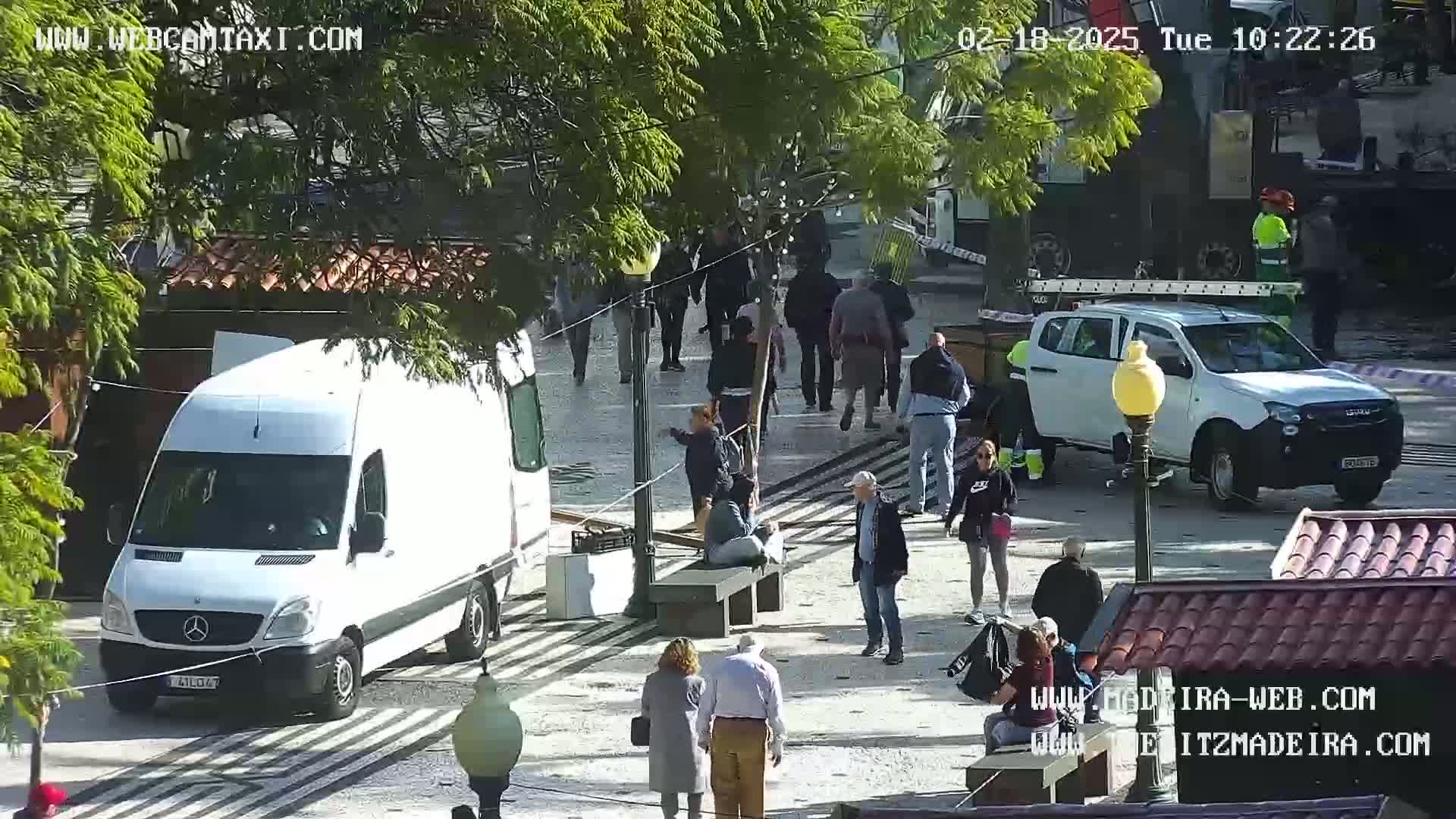 A sunny outdoor scene shows a group of people standing and sitting around a paved area with several vehicles, including a white van and a white pickup truck.