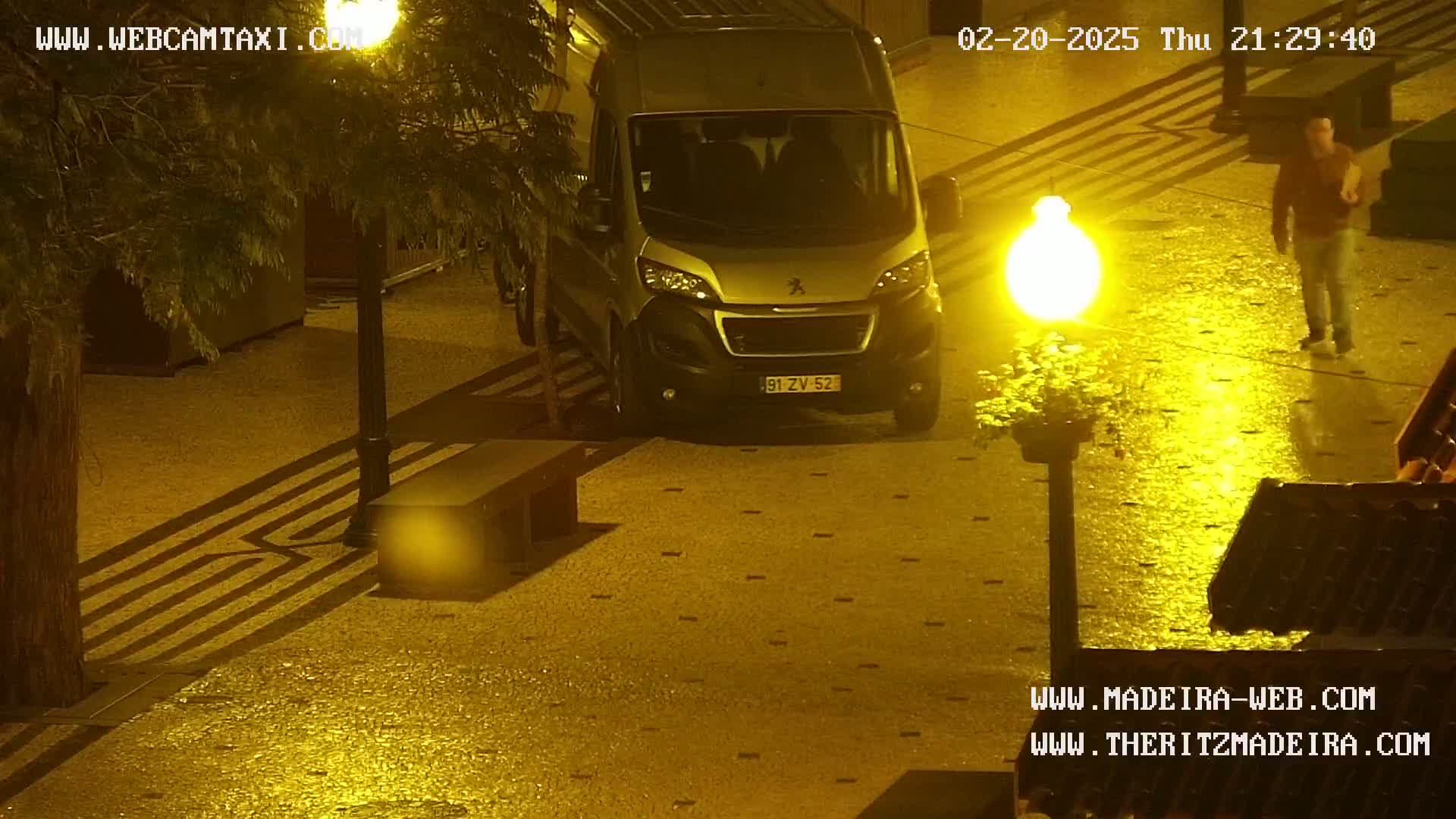 A silver van is parked in a wet, dimly lit paved area at night, near a person walking and a streetlamp.
