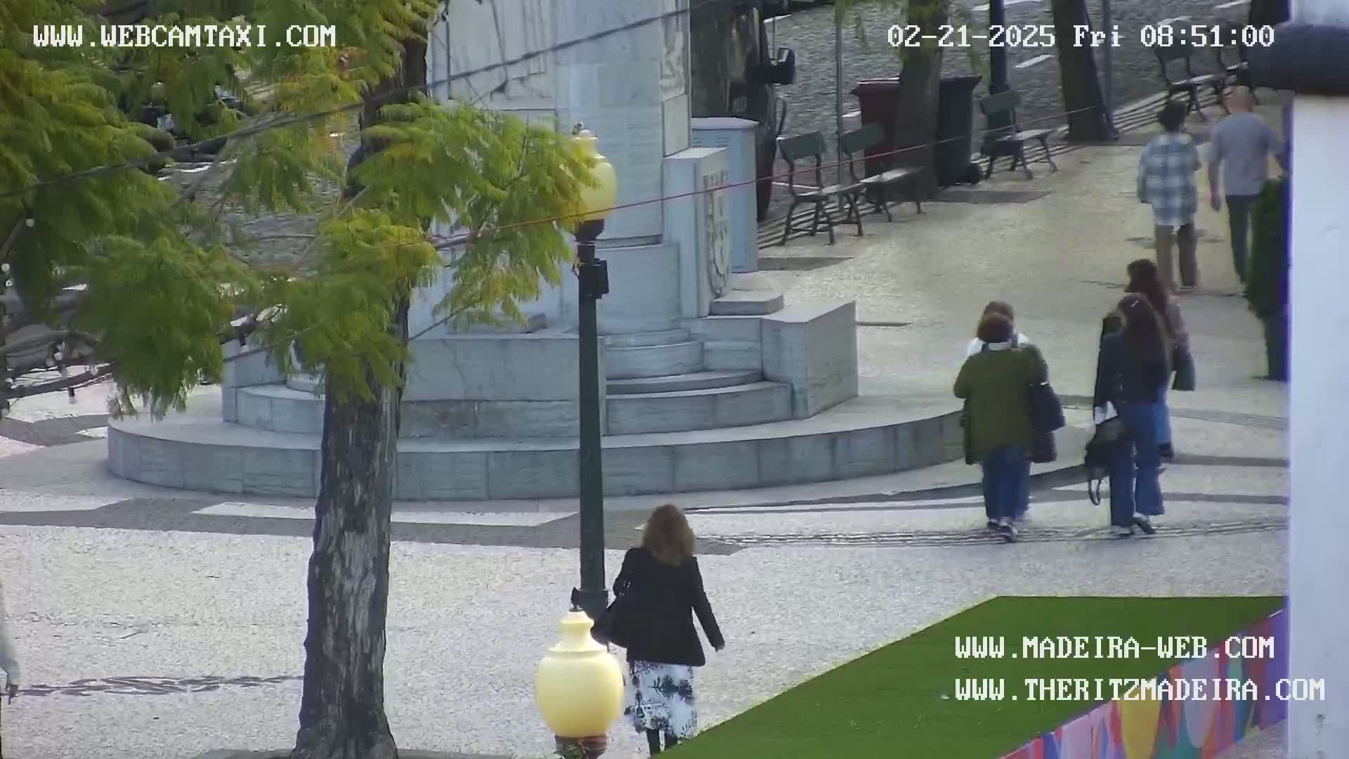 A woman walks near a stone monument in a plaza under a partly cloudy sky.