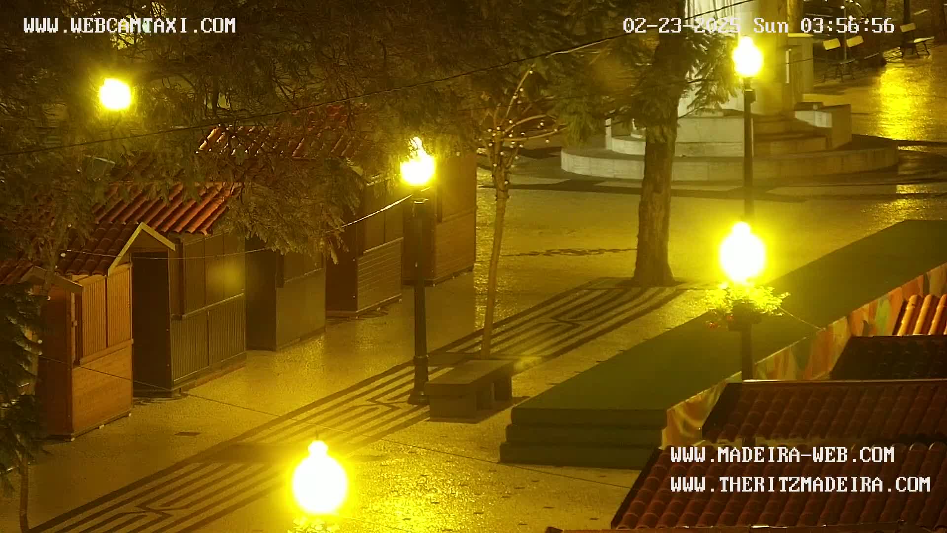 A wet, nighttime scene shows several small wooden structures illuminated by bright streetlights in a paved plaza.