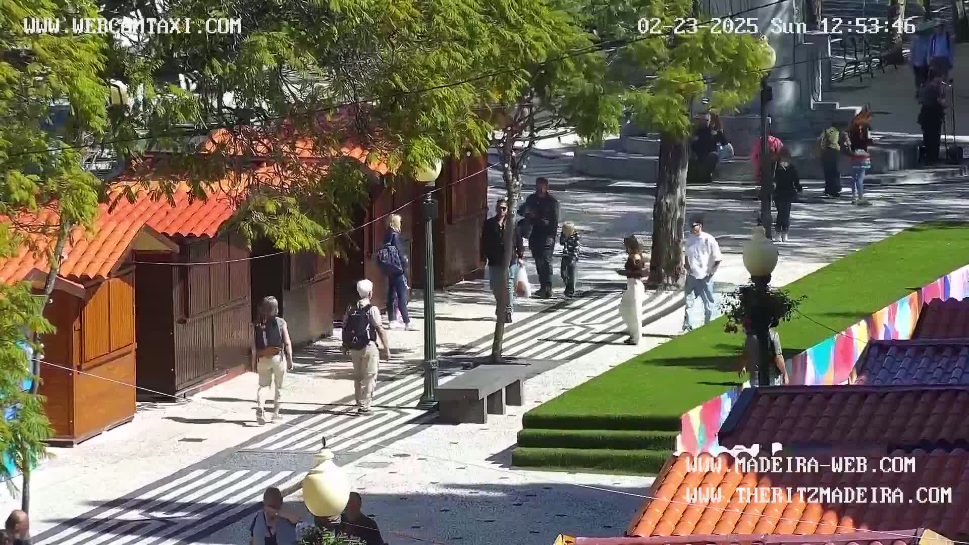 A sunny outdoor plaza scene shows several people walking past small wooden kiosks with orange roofs and a grassy area with a patterned walkway.