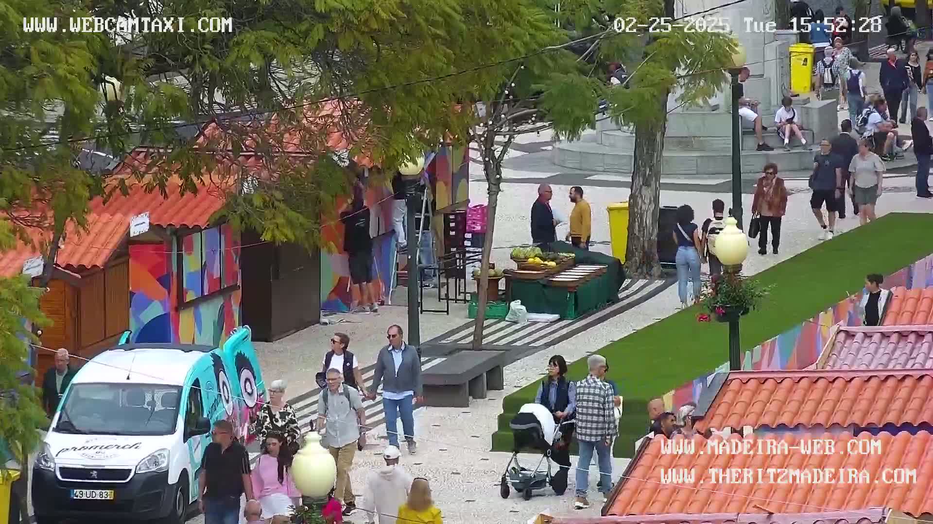 A partly cloudy day shows a paved outdoor market area with colorful stalls, people walking around, and a van parked nearby.