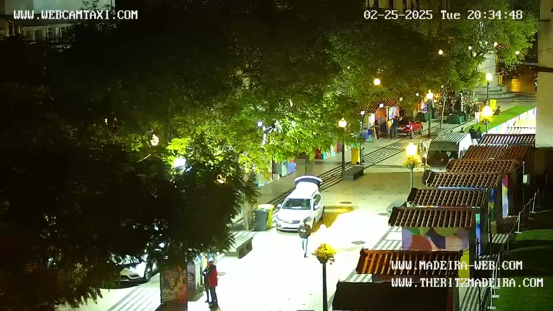 An aerial nighttime view of a paved pedestrian area with several small market stalls, some cars, and pedestrians, illuminated by streetlights under mostly clear skies.
