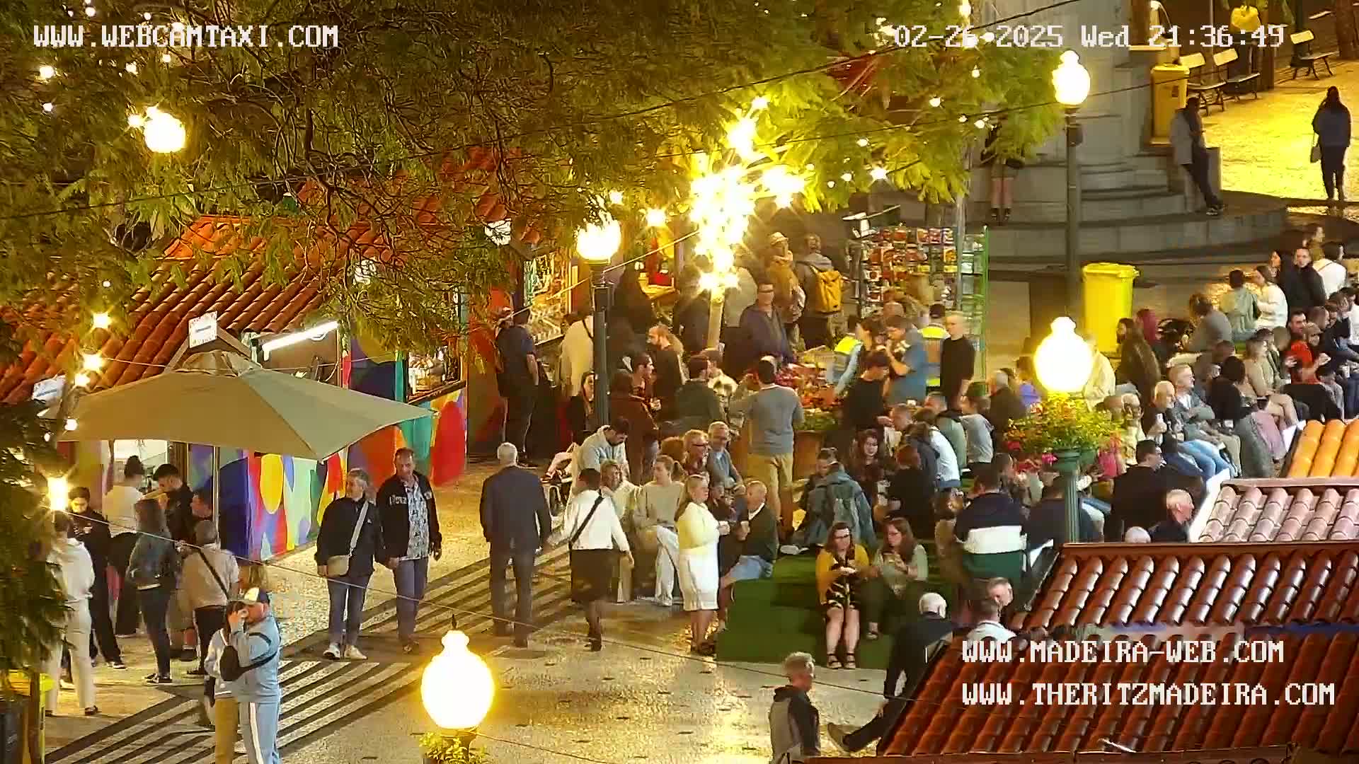 A nighttime outdoor scene shows a large crowd of people gathered around market stalls, illuminated by string lights and street lamps.