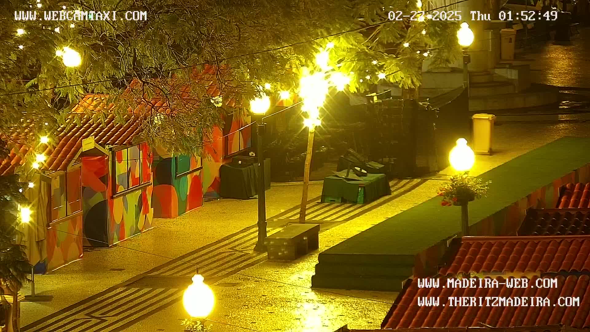 A nighttime scene of a paved plaza with colorful, decorated stalls illuminated by string lights and street lamps, under wet conditions.