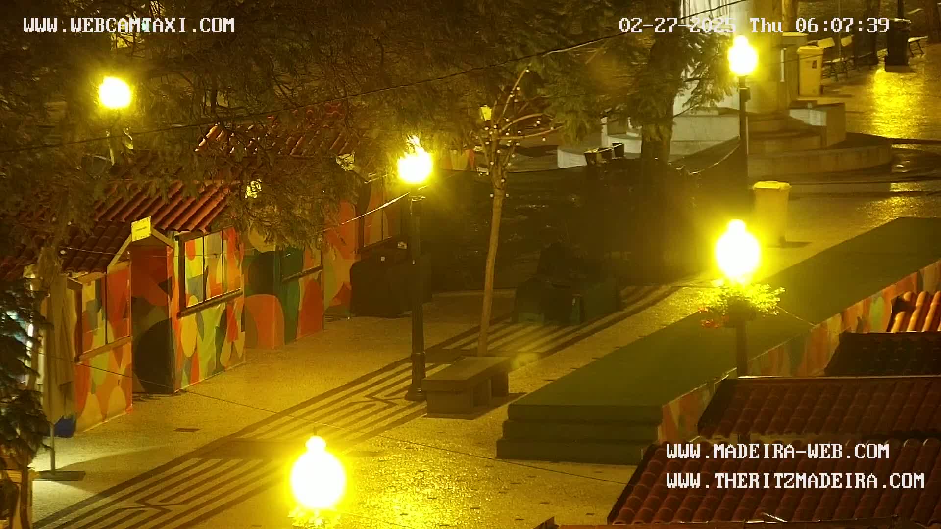 A nighttime view of a wet, brightly lit plaza with colorful buildings and streetlights.