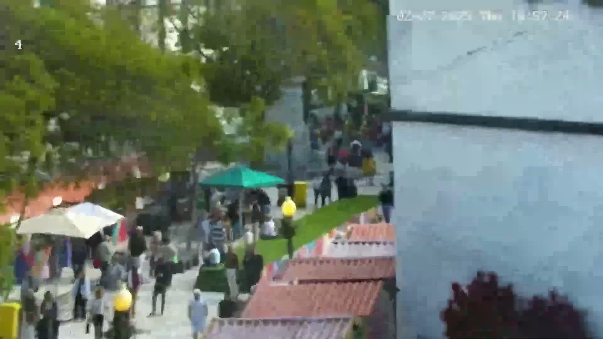 A large crowd of people gathers outdoors on a mostly sunny day amidst numerous market stalls and green trees.