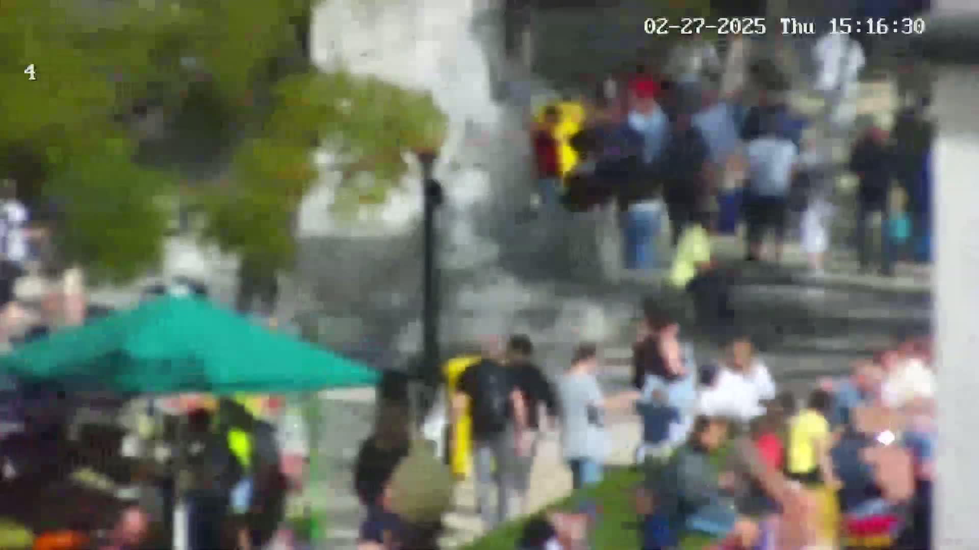 A large, blurred crowd of people gathers outdoors on a partly sunny day, near a green canopy and a stone structure.
