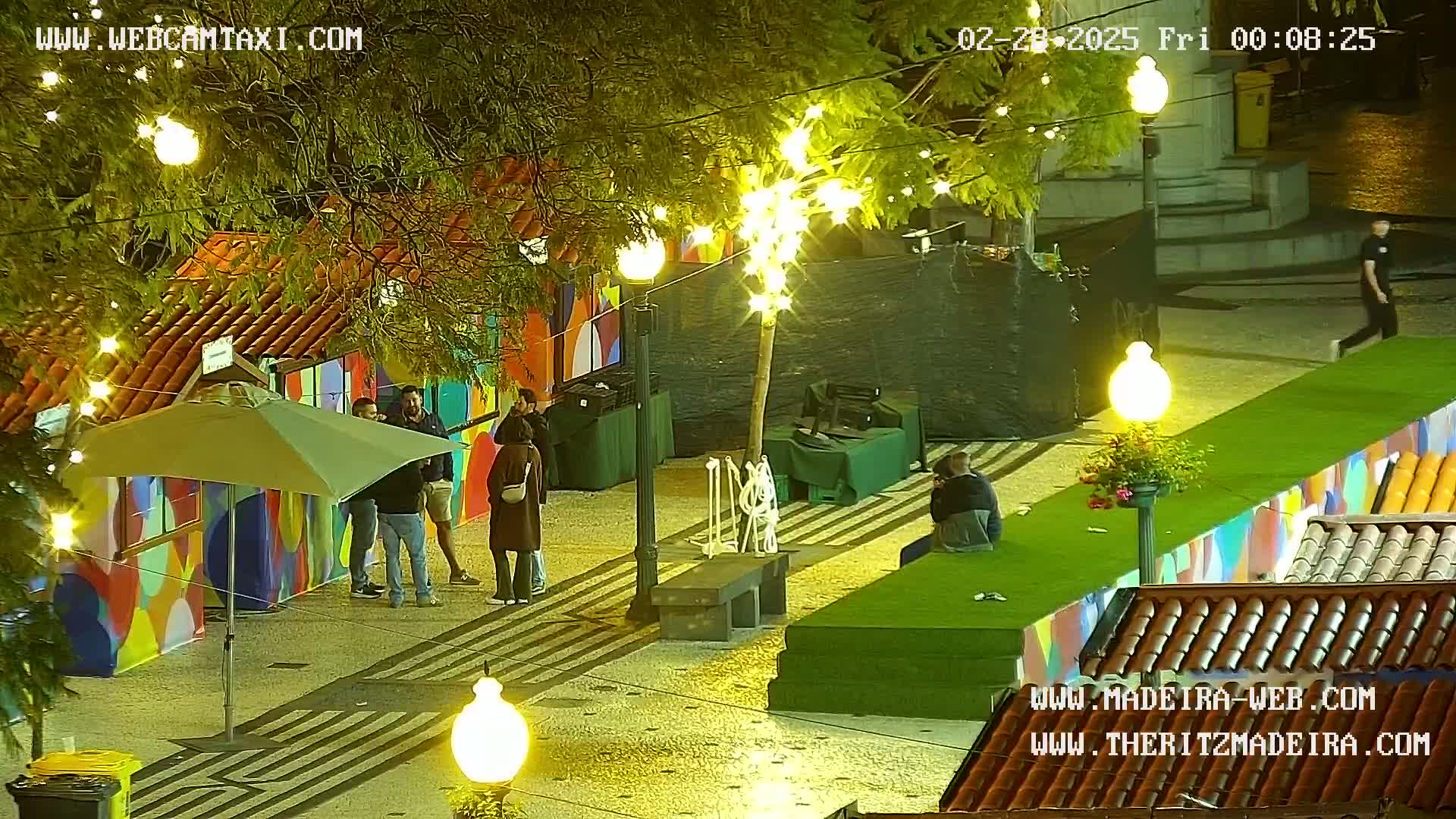 At night, under string lights, several people stand and sit near brightly colored structures and a green artificial lawn, all illuminated by streetlights.