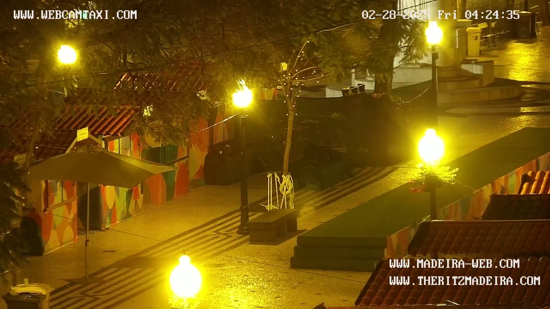 A nighttime outdoor scene shows a paved area with brightly lit street lamps, colorful buildings, and a small umbrella-covered stand, under a light rain.
