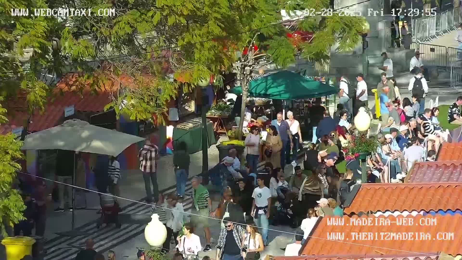 A sunny outdoor market scene with numerous people milling around stalls and under umbrellas.