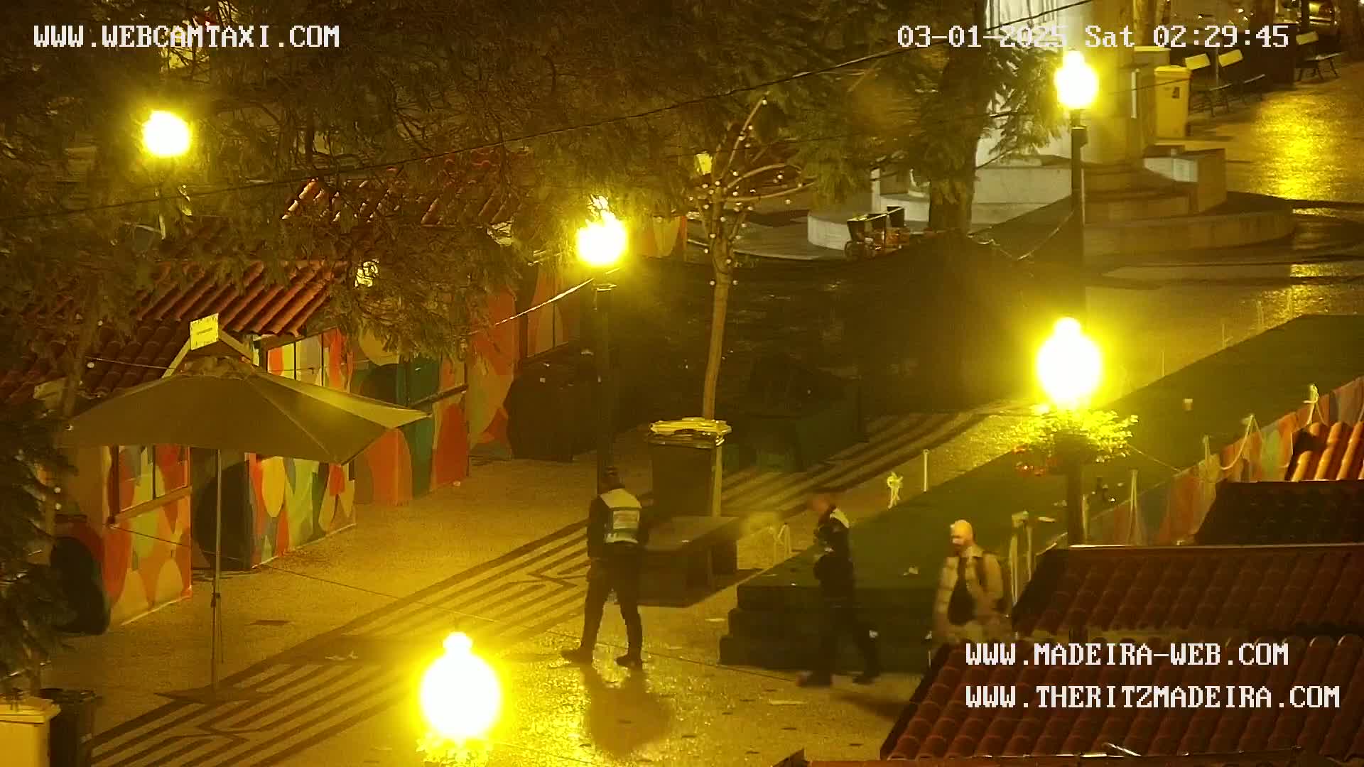 A nighttime, wet outdoor scene shows several people standing near brightly lit stalls and streetlamps in a paved area.