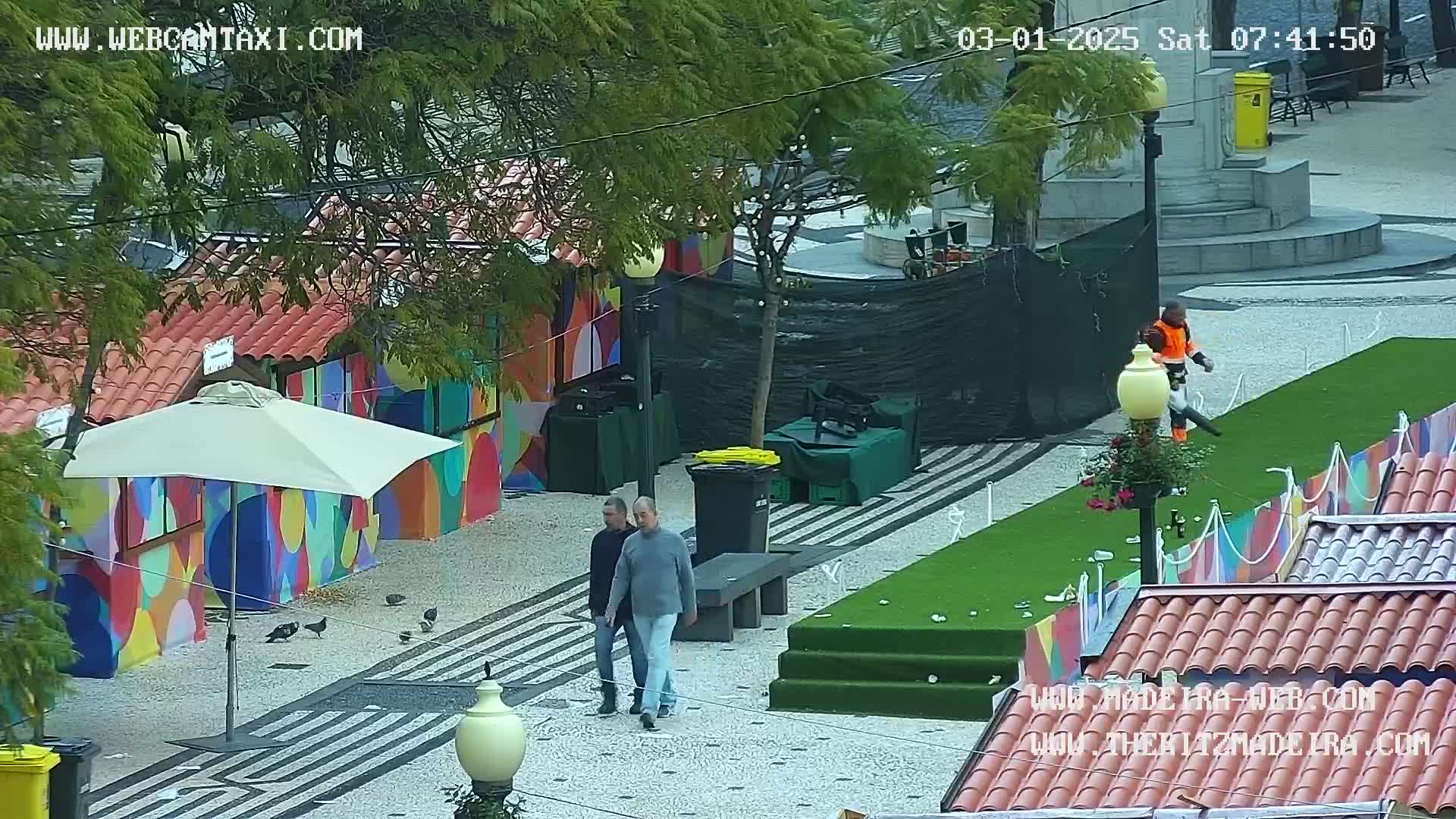 Two men walk past colorful stalls and a patch of artificial turf in a plaza, where a person is using a leaf blower.