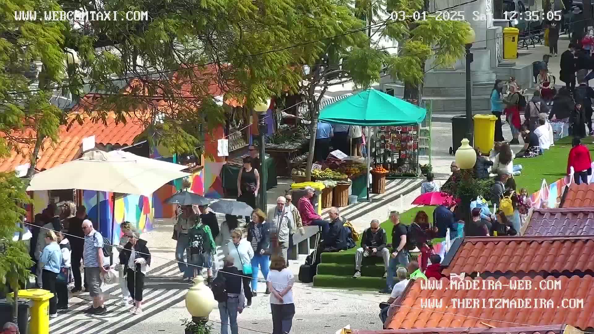 A sunny outdoor market scene shows numerous people gathered around colorful stalls and under umbrellas, with some sitting on grassy areas.