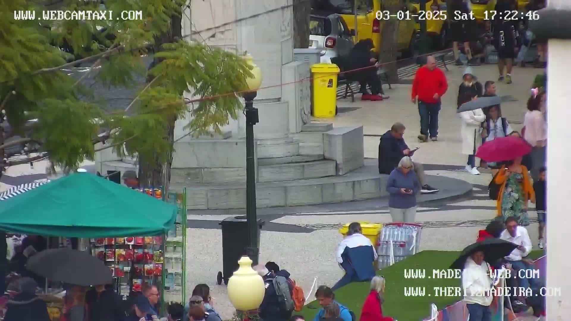 A rainy outdoor plaza scene shows numerous people walking, sitting, and using umbrellas; some are near a vendor's stall under a green awning.