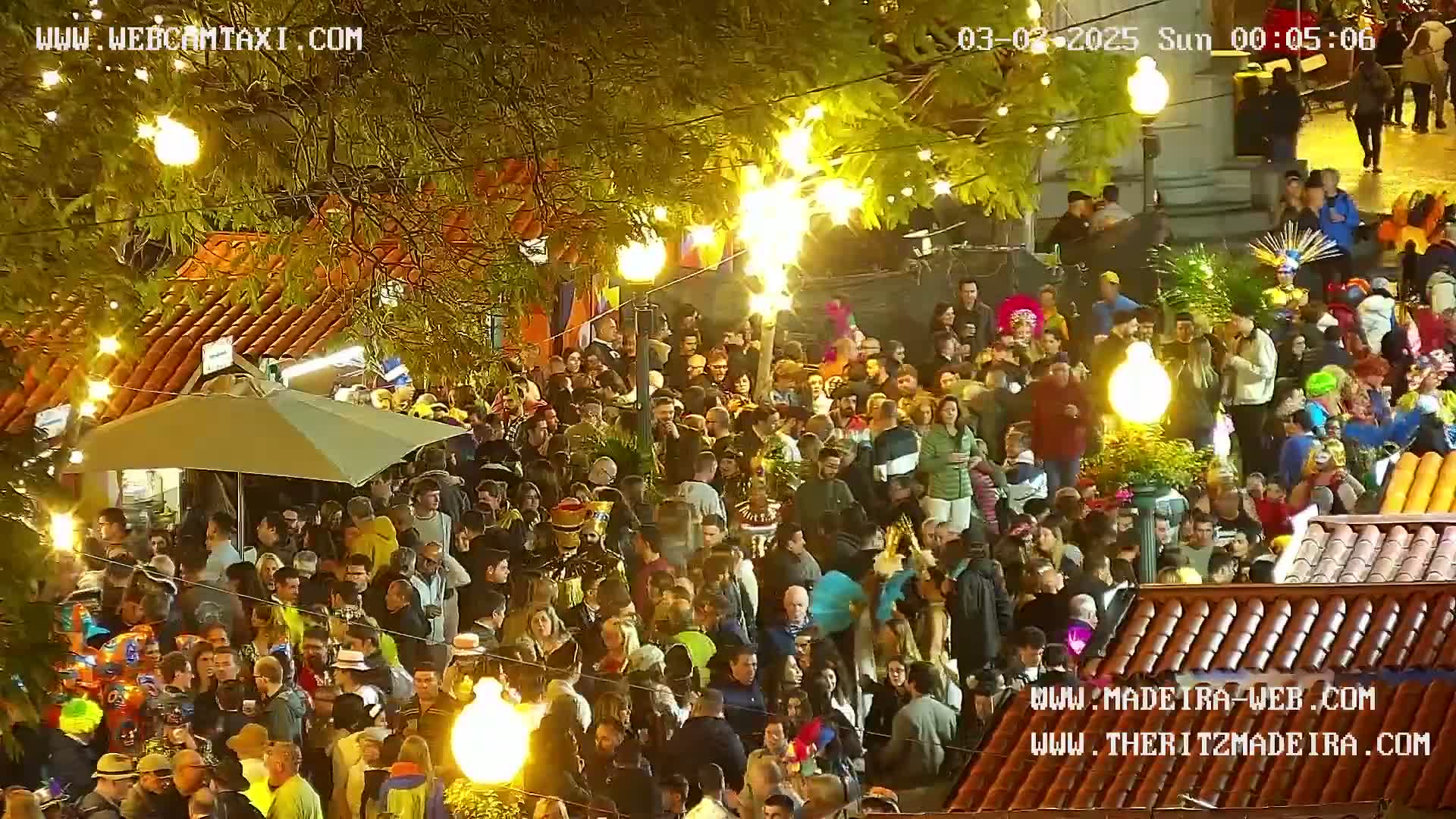 A large crowd of people, many in costumes, gathers outdoors at night under trees with string lights, illuminated by numerous lights.