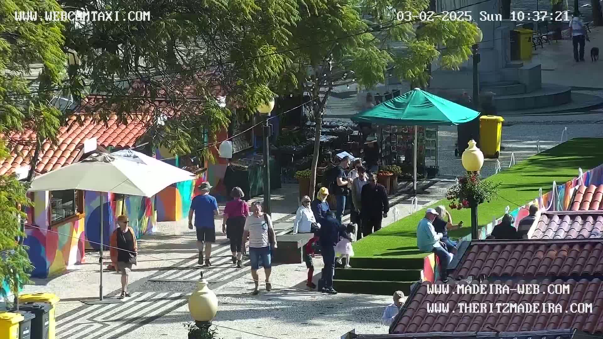 A sunny outdoor market scene shows people walking and shopping amongst colorful stalls and small green lawns.