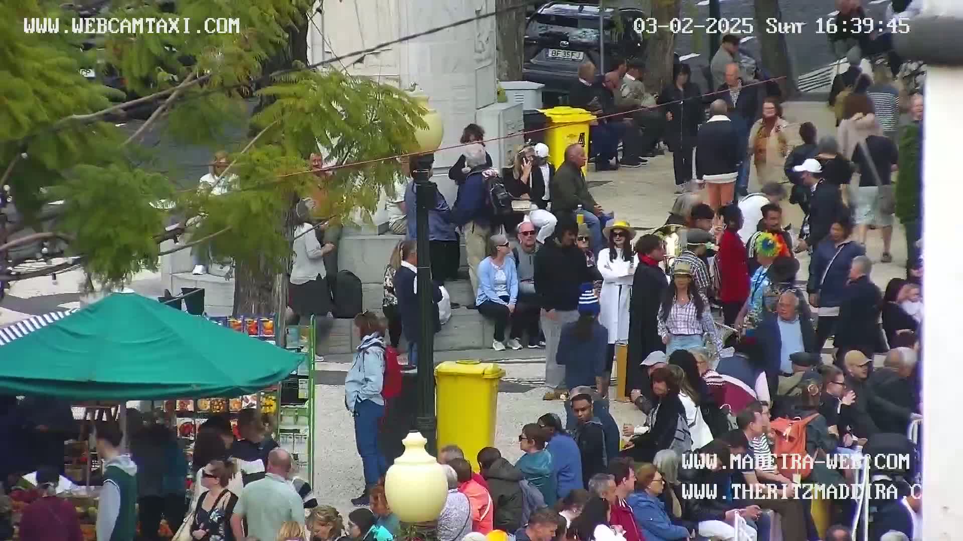 A large crowd of people gathers in a sunny outdoor plaza, some sitting and others standing.