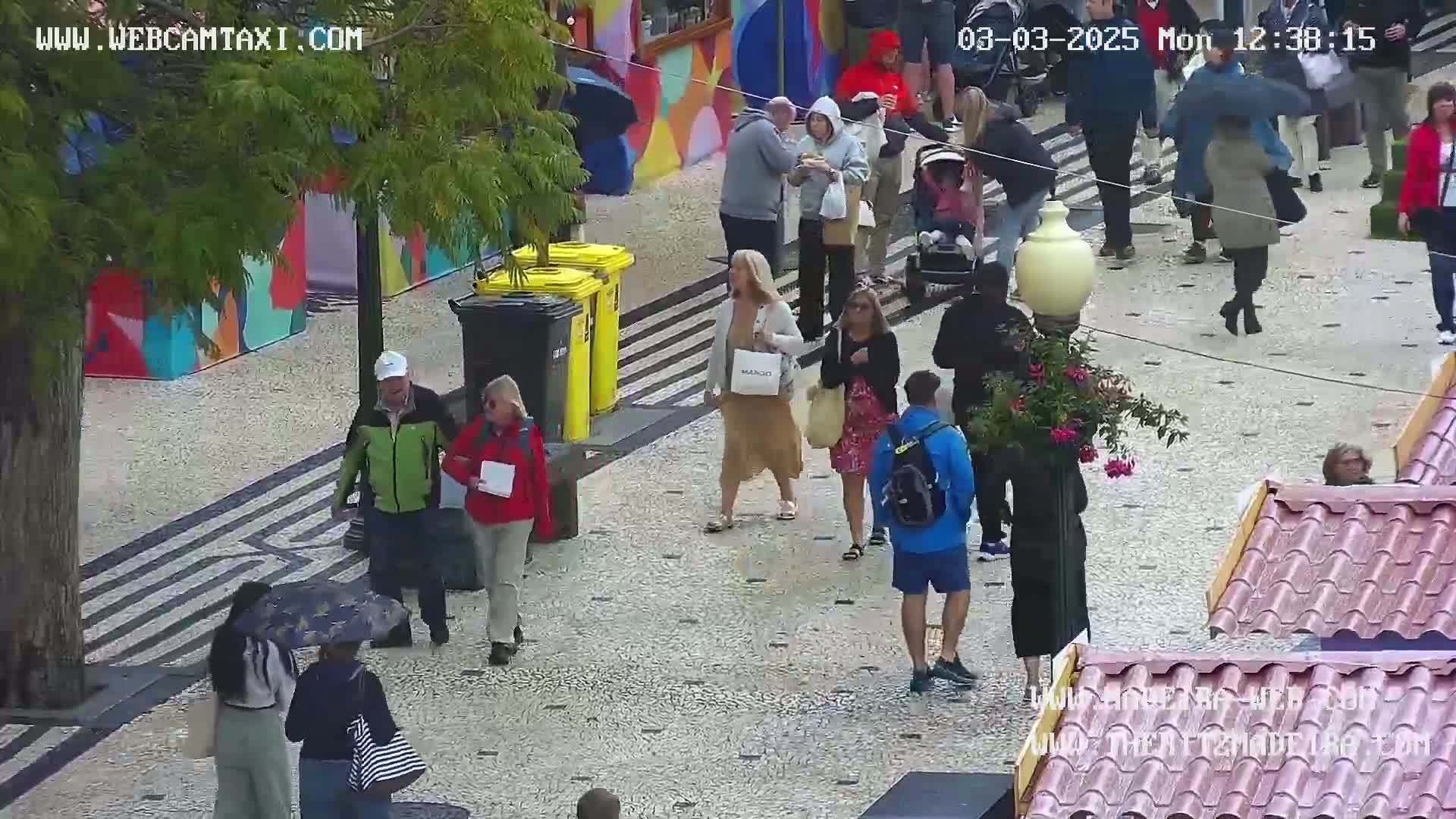 A paved pedestrian area with several people walking around, some carrying shopping bags and umbrellas under a lightly overcast sky.
