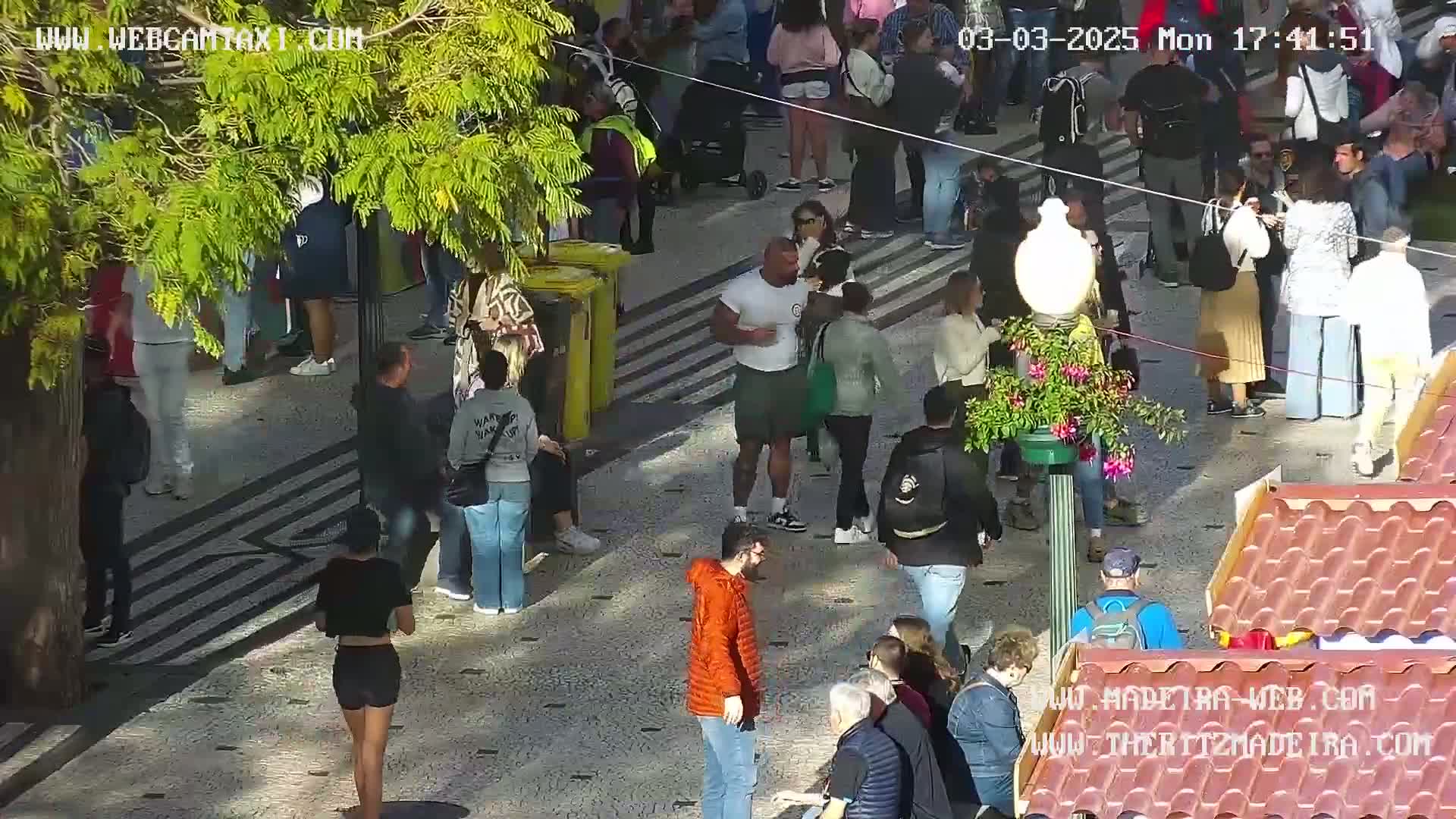 A sunny outdoor scene shows many people walking on a paved pedestrian area, some near a yellow bin and others near market stalls.