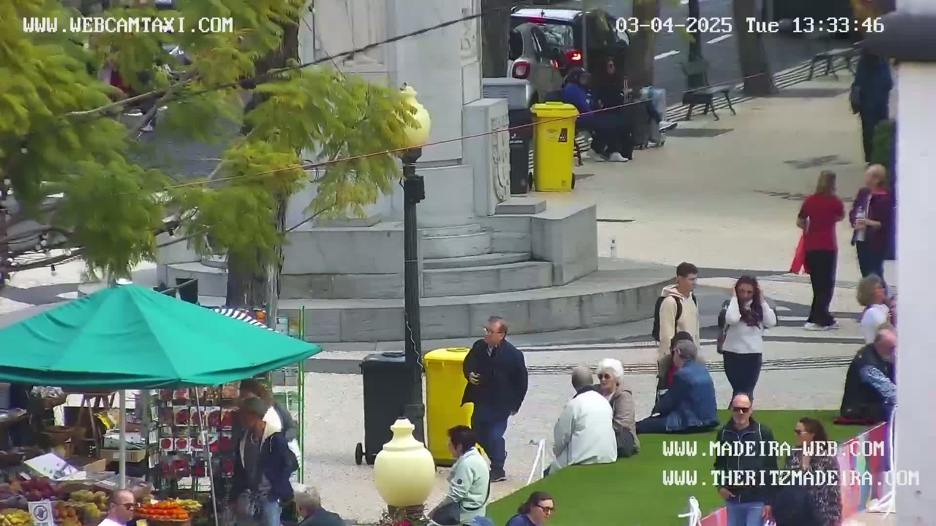 A sunny outdoor plaza scene shows people walking, sitting, and standing around a market stall under a green umbrella, near a stone monument and yellow trash cans.