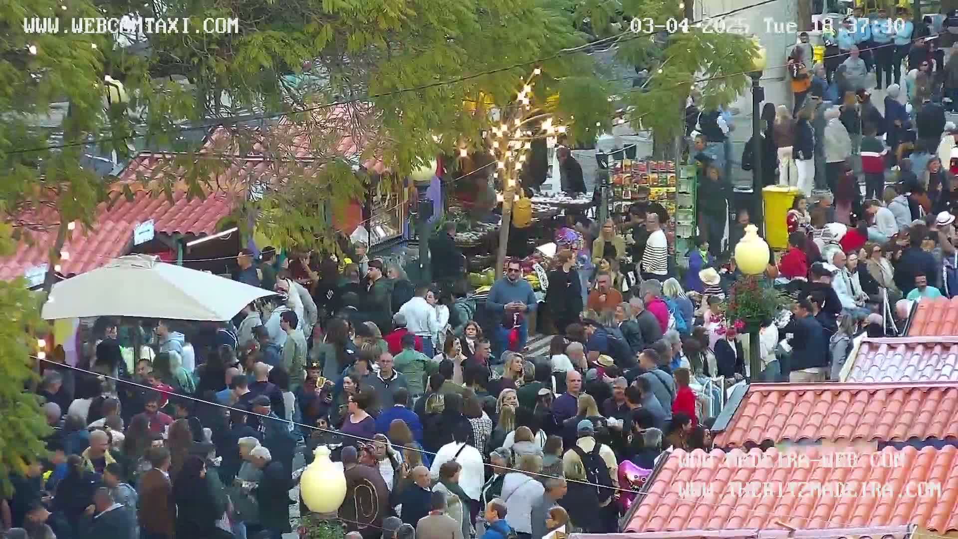 A large crowd of people gathers outdoors in an open-air market under a partly cloudy sky.