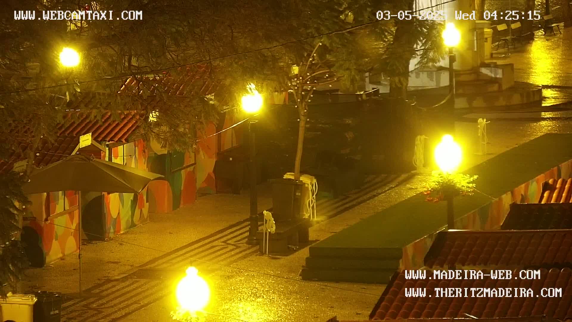 A rain-slicked, brightly lit plaza at night features colorful buildings and several streetlights casting a warm yellow glow.