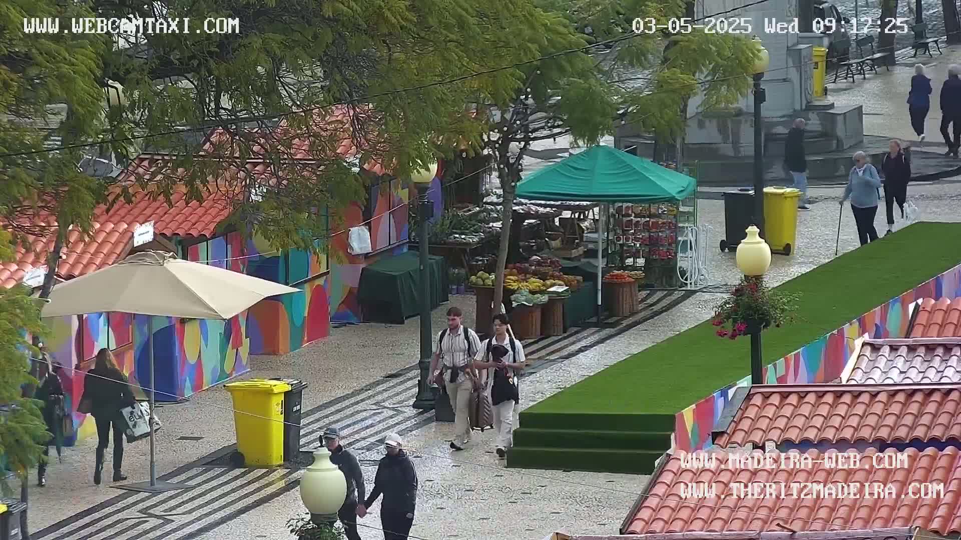 A paved outdoor market area with colorful buildings, a green artificial turf section, several people walking around, a fruit stand under a green canopy, and an umbrella-covered table.  The weather appears overcast.