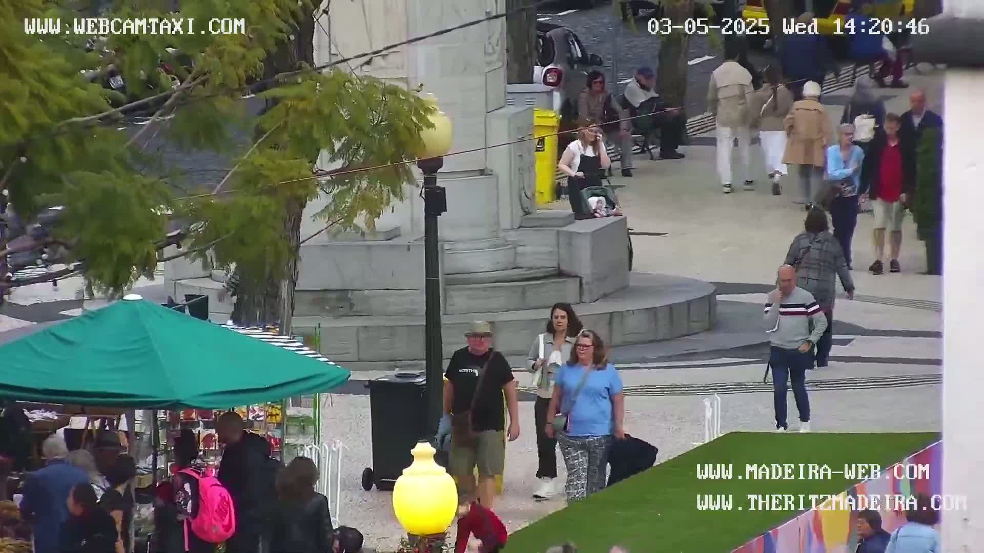 A mostly sunny outdoor scene shows a paved plaza with many people walking around, some near a green market umbrella and others near a stone monument.