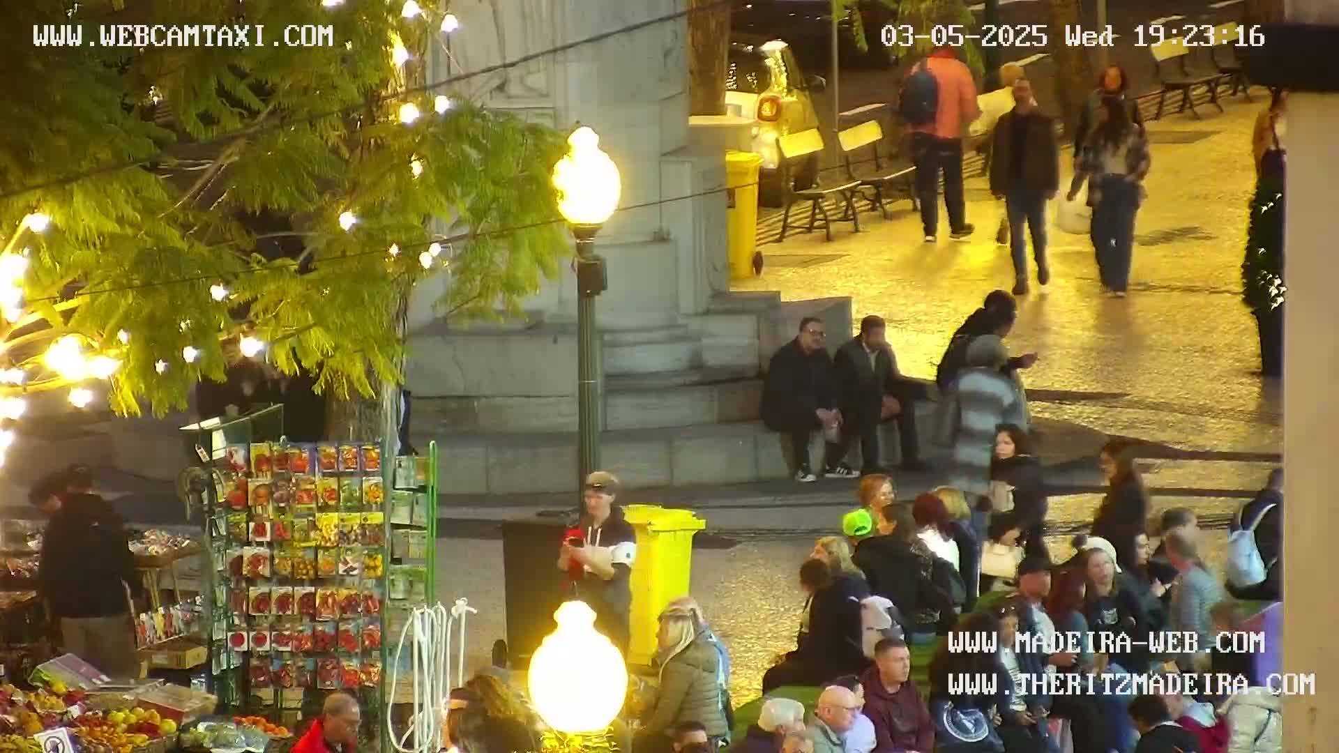 A nighttime outdoor scene shows a city square filled with people, some sitting and some standing, near a monument and a market stall under a tree with string lights.