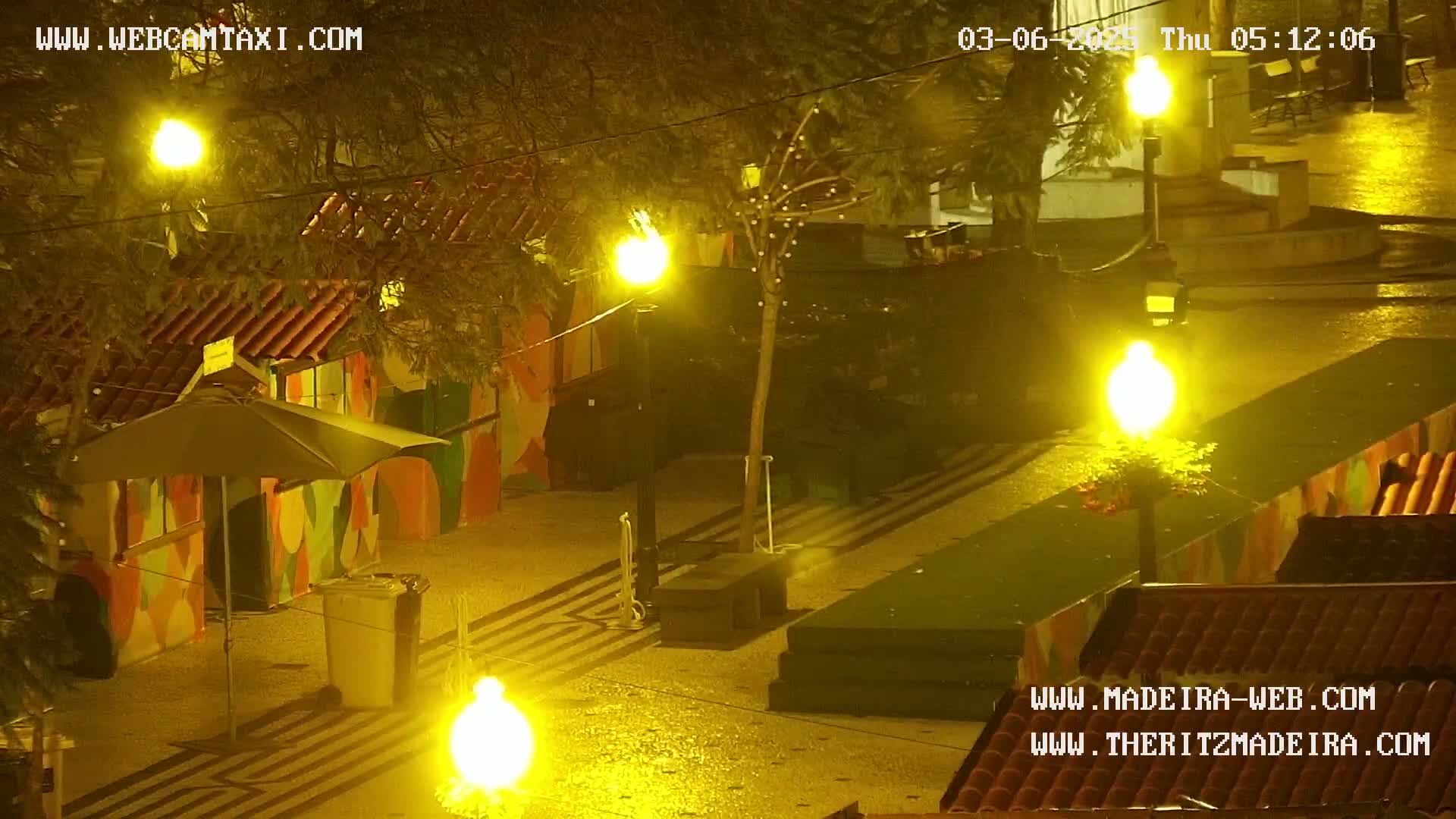 A nighttime outdoor scene shows a wet paved area with brightly lit street lamps, colorful buildings, and a small umbrella.