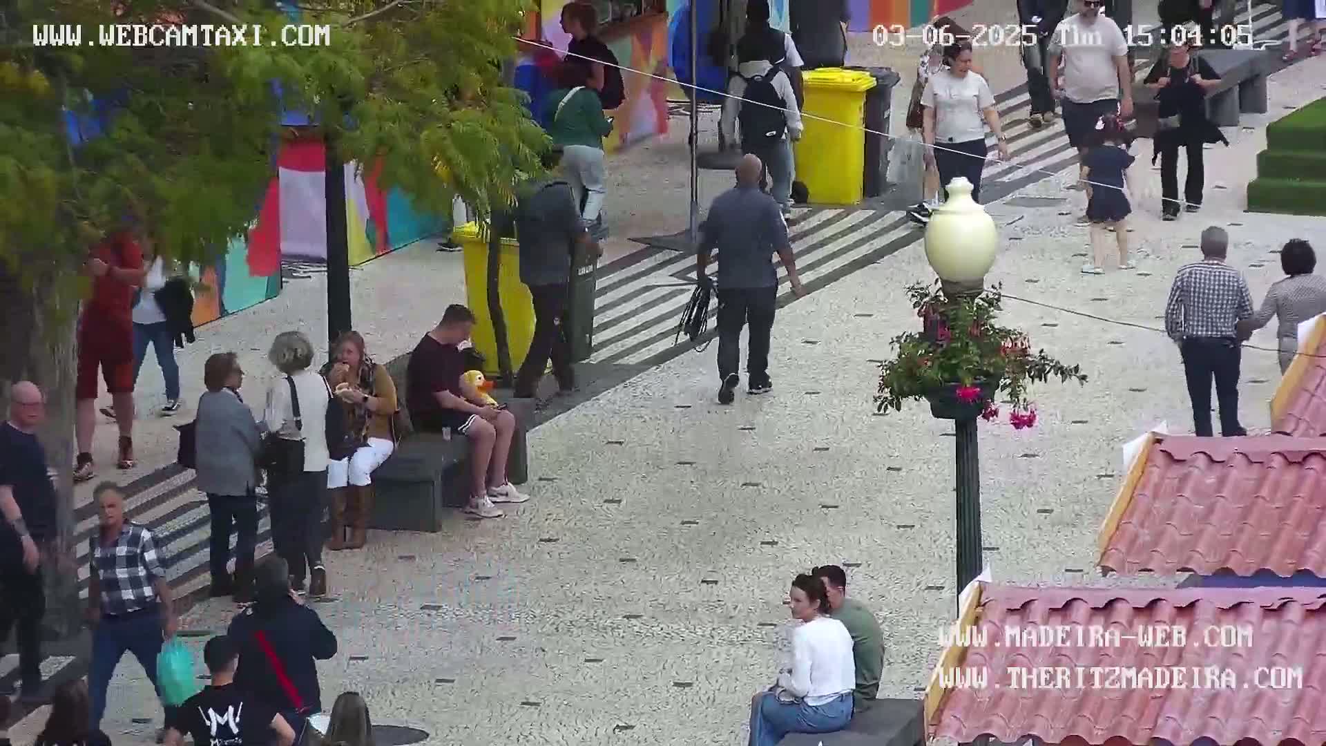 A paved pedestrian area with numerous people walking and sitting, some in groups, under a partly cloudy sky.