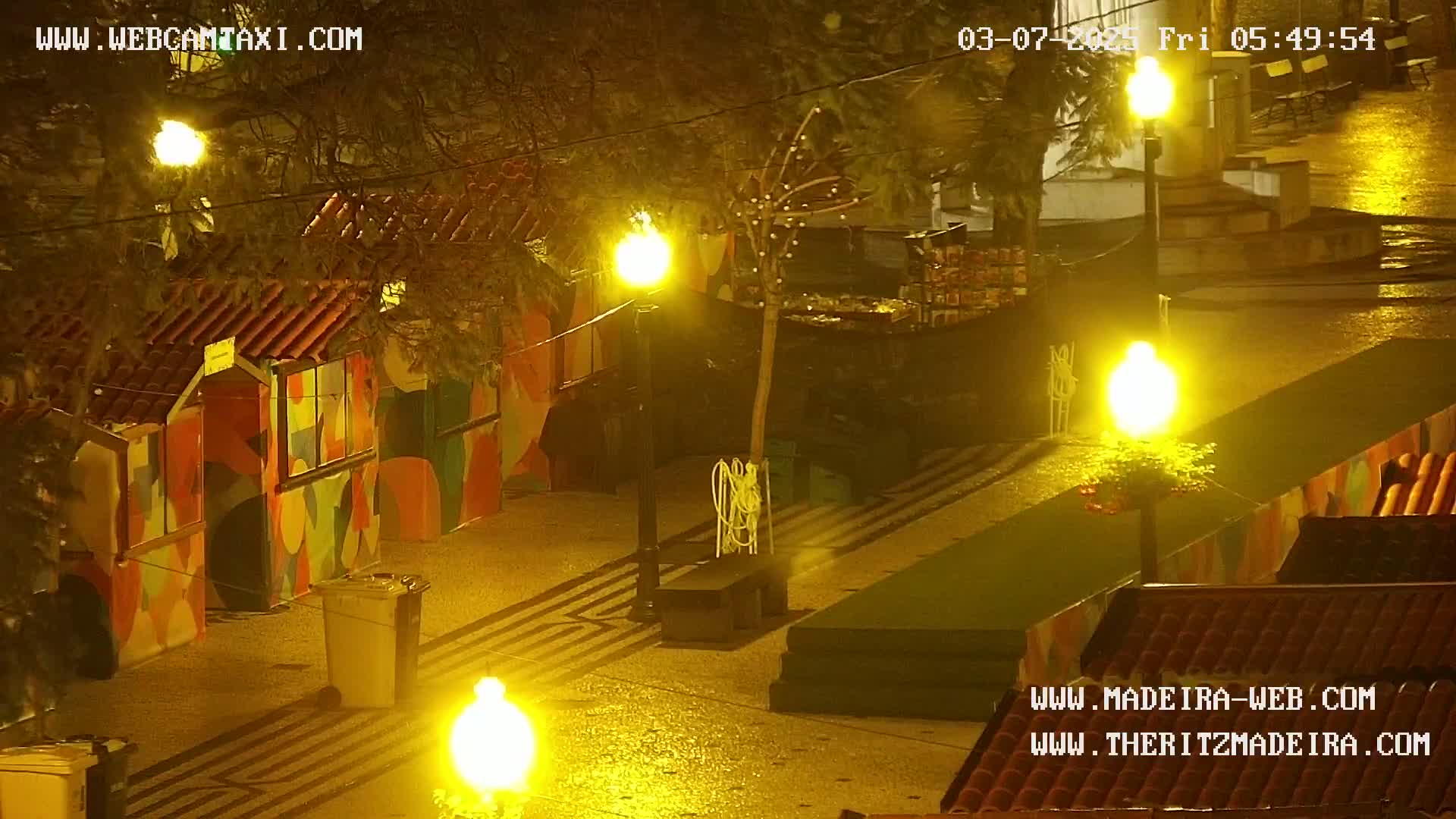A nighttime view of a wet, brightly lit plaza with colorful stalls and several streetlights.