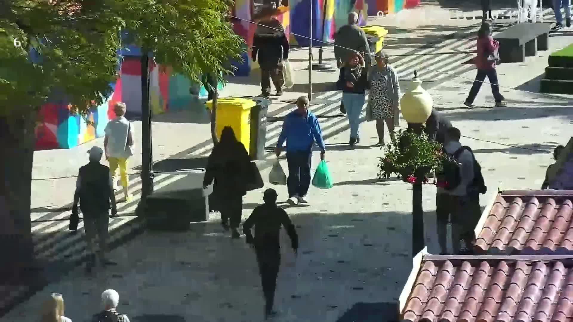 A sunny day shows numerous pedestrians walking on a paved pathway, some carrying shopping bags, past colorful buildings and trash cans.