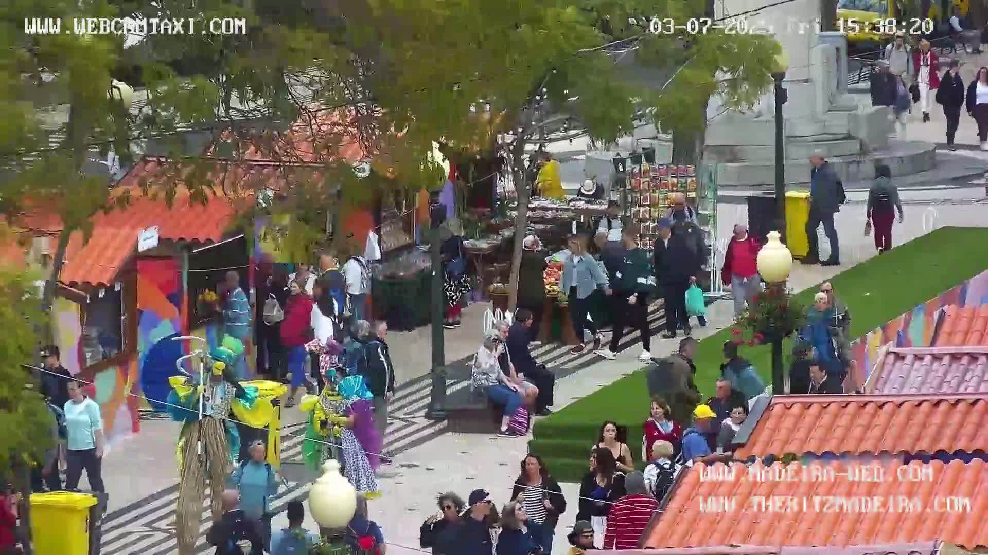 A bustling outdoor market scene with numerous people browsing colorful stalls under a sunny sky.
