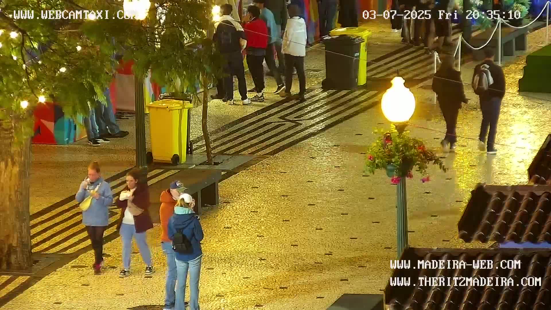 A wet, paved pedestrian area at night is sparsely populated with several people walking and standing around yellow trash cans and under streetlights.